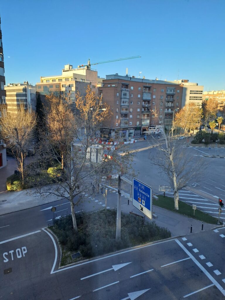 A cityscape view of Madrid with residential and commercial buildings, leafless trees, a road intersection with traffic signs, and a construction crane in the background under a clear blue sky.