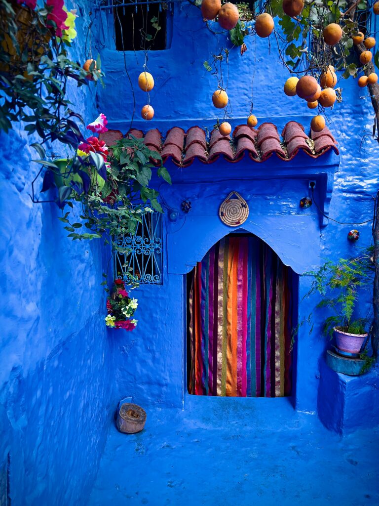 Traditional blue-washed house in the medina of Chefchaouen, Morocco, with ornate doorways and vibrant flower pots adorning the walls.